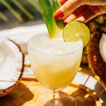 a light yellow spritz being garnished with pineapple leaves and a lime wedge