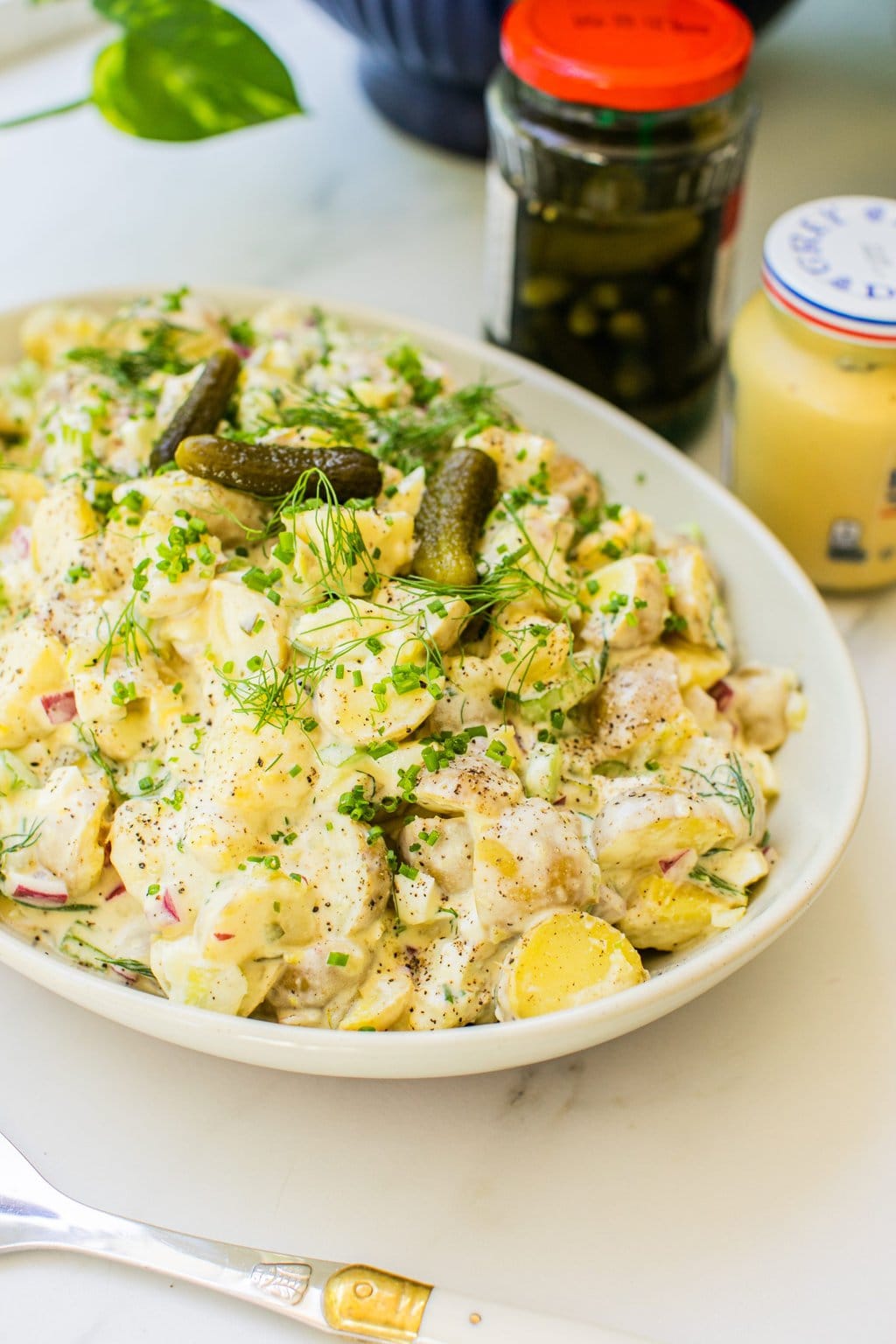 a white bowl of potato salad topped with fresh dill and dill pickles, as well as a jar of pickles and dijon mustard out of focus in the background