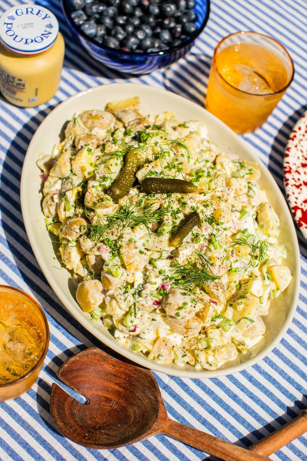 a blue and white striped table cloth with a large cream colored serving bowl on top, filled with dill potato salad. 