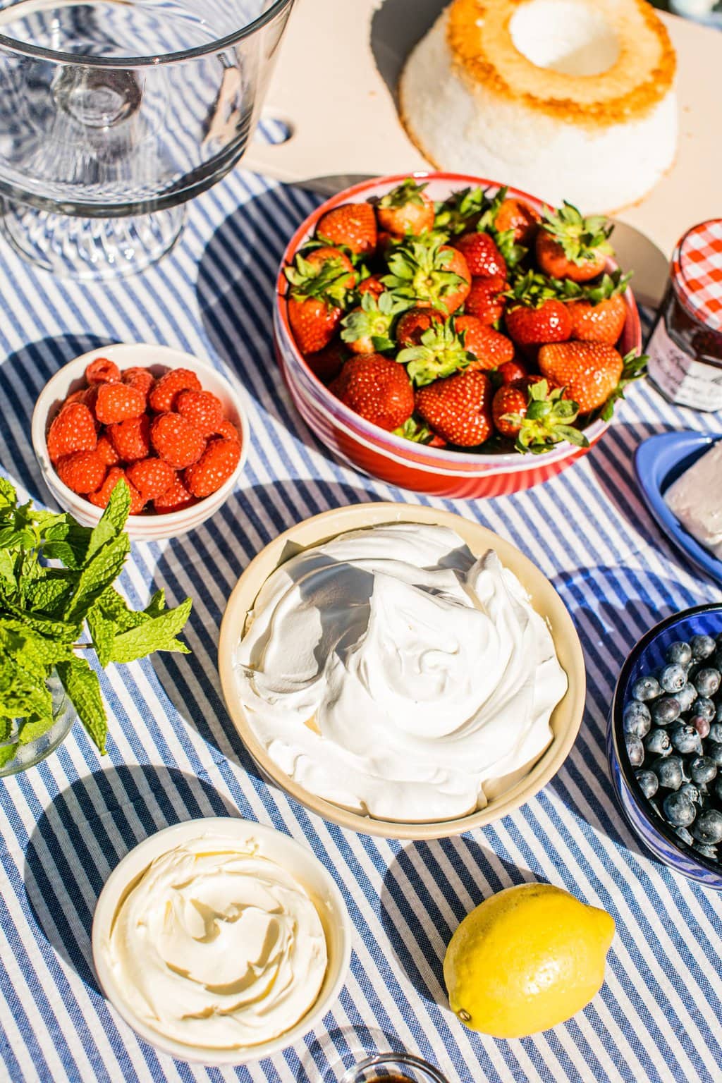 bowls of strawberries, raspberries, blueberries, jam, mascarpone cheese, cream cheese, lemon, pound cake, and mint on a blue and white striped cloth