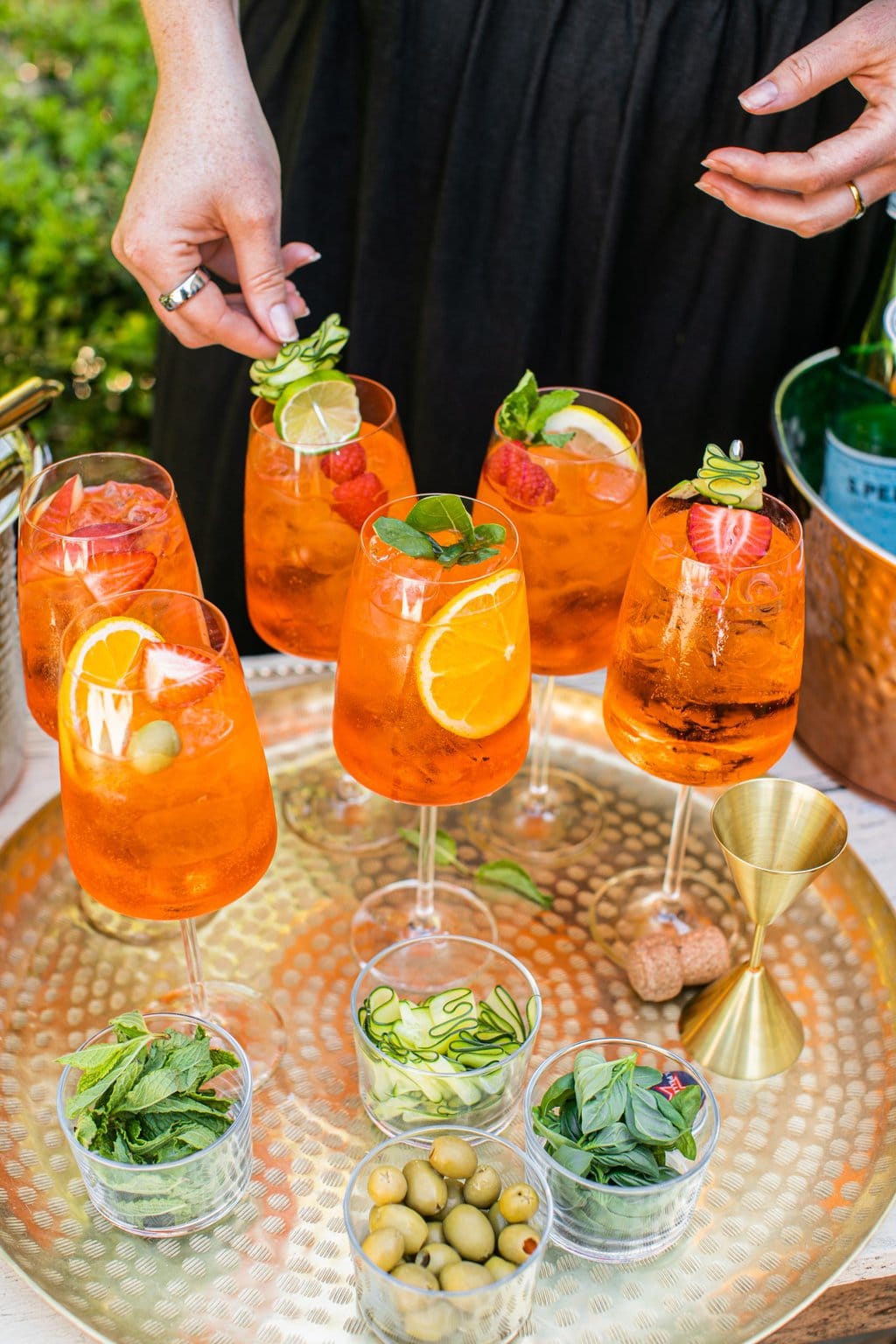 a gold platter with 6 glasses of aperol spritzes garnished with different things