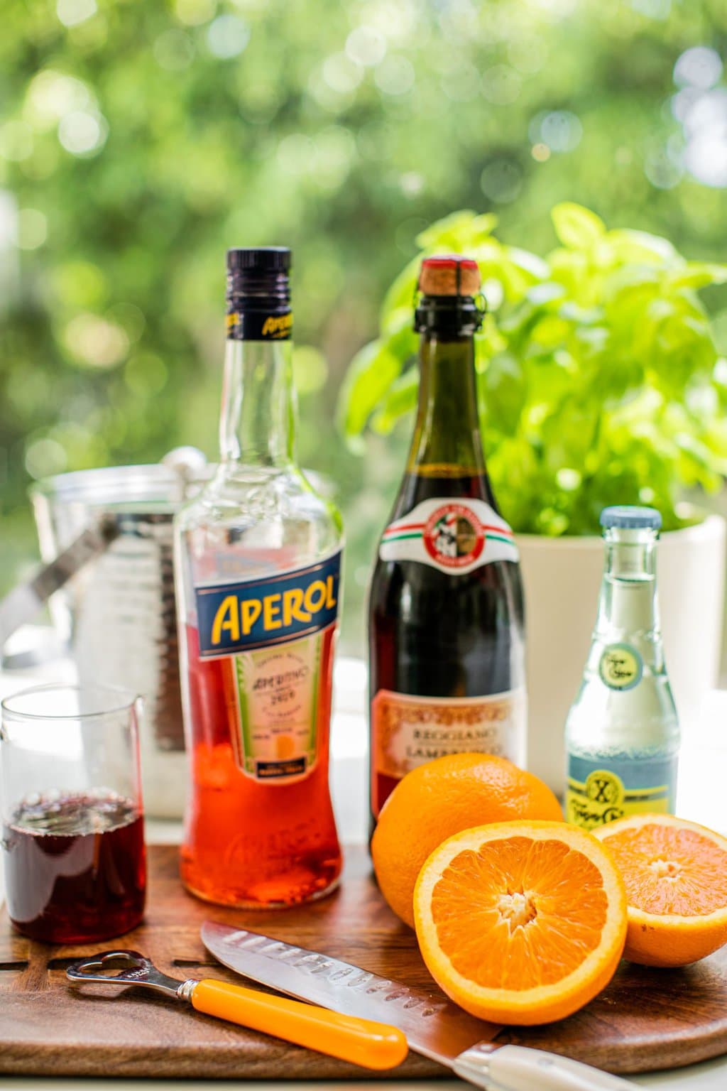 aperol, lambrusco, topo chico, oranges, and pomegranate juice on a wooden chopping board beside a knife and bottle opener