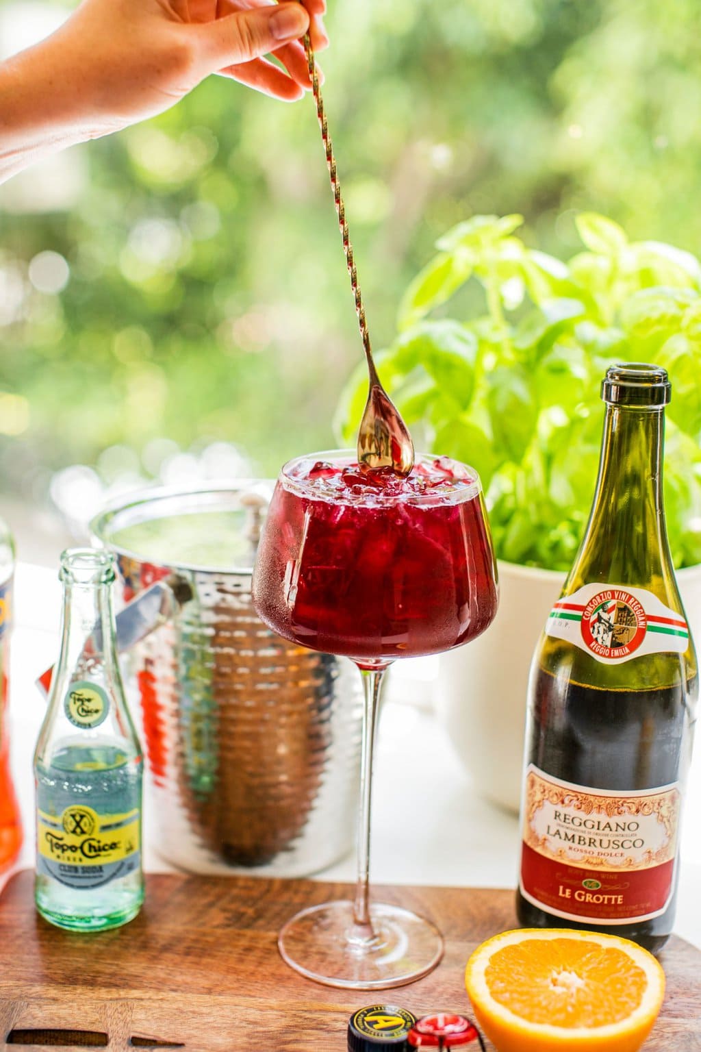 a spritz in the middle of ingredients like lambrusco, topo chico, and an orange, being mixed by a long golden cocktail stirrer