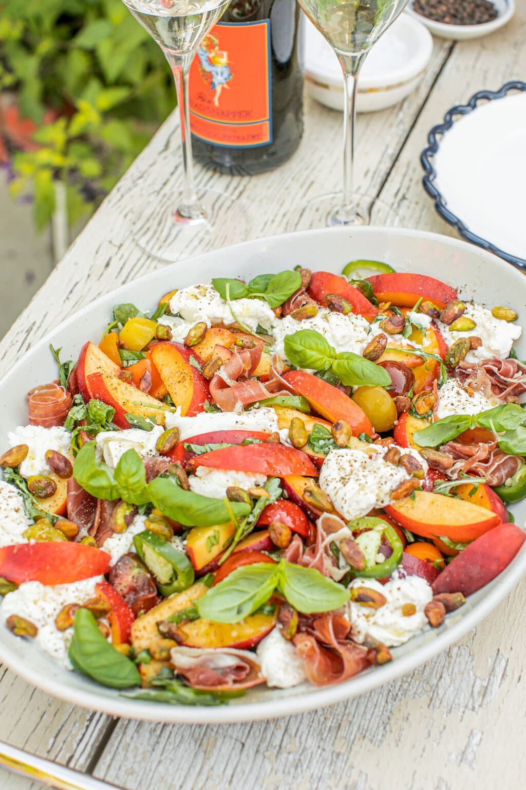 a bowl full of burrata salad with peaches sitting on a white washed wooden table with a wine bottle and glasses in the background