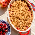 peach crisp in a large baking dish beside a bowl of fresh cherries and whole peaches