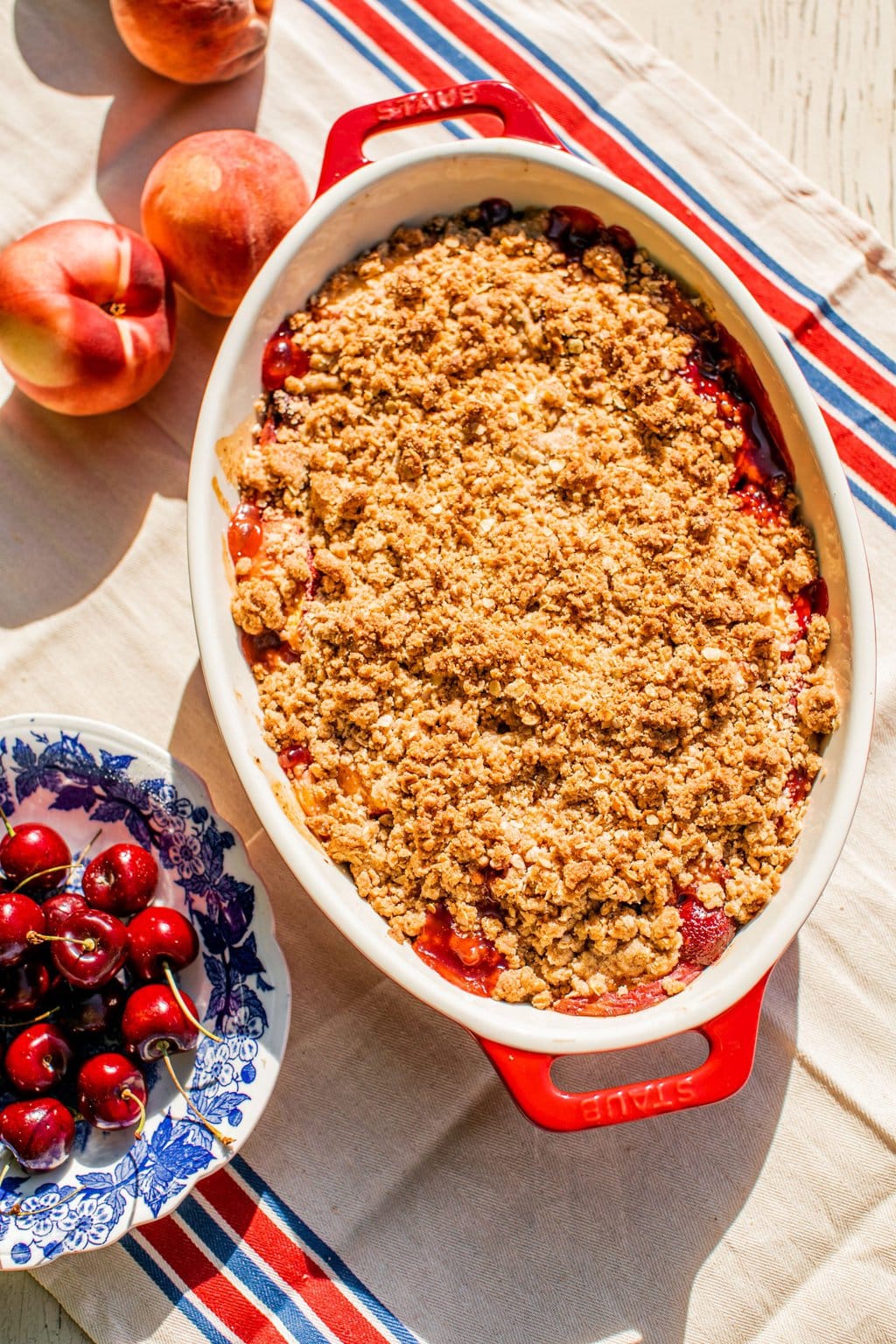 peach crisp in a large baking dish beside a bowl of fresh cherries and whole peaches