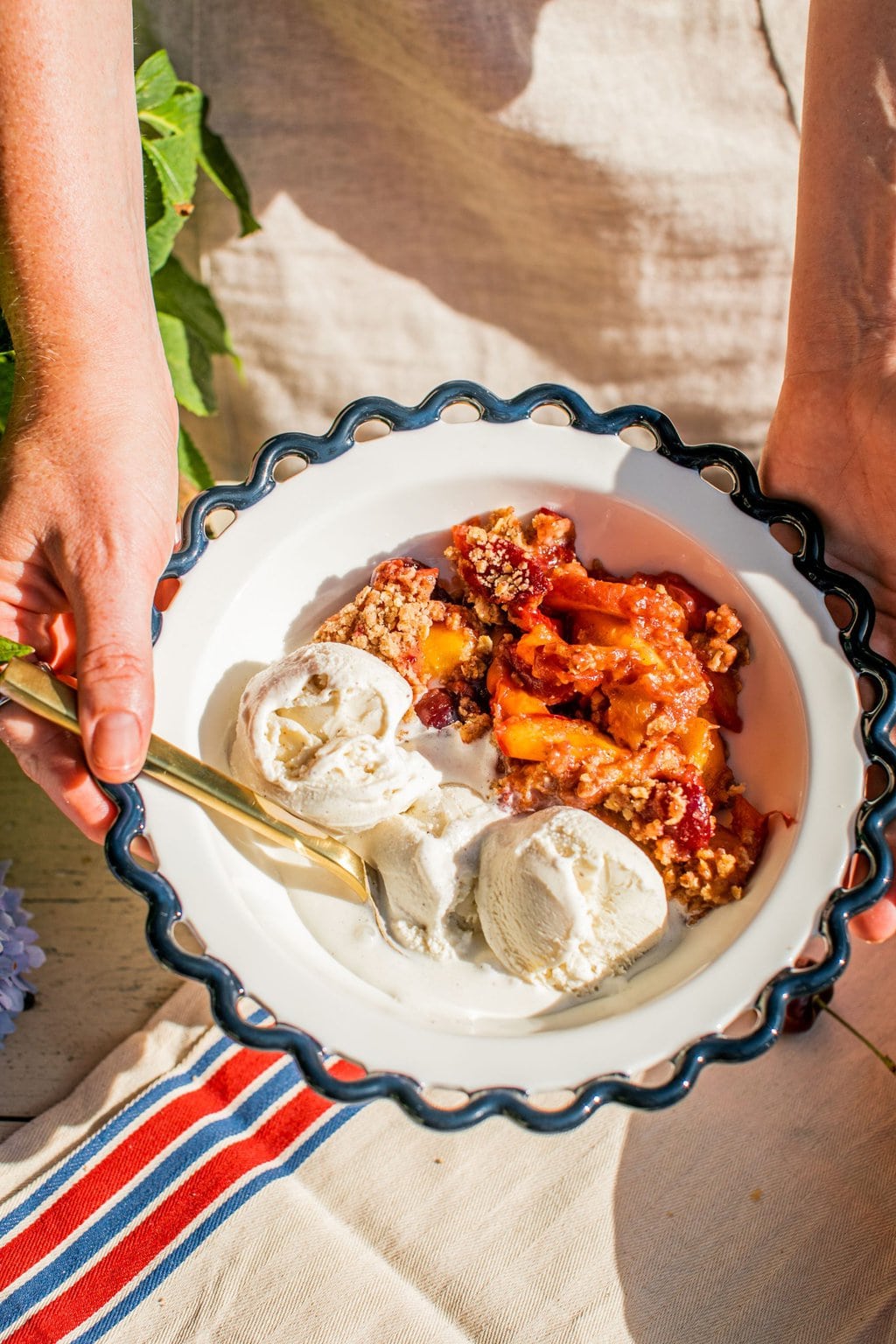 two hands holding a bowl of a fruity crisp dessert alongside a few scoops of vanilla ice cream