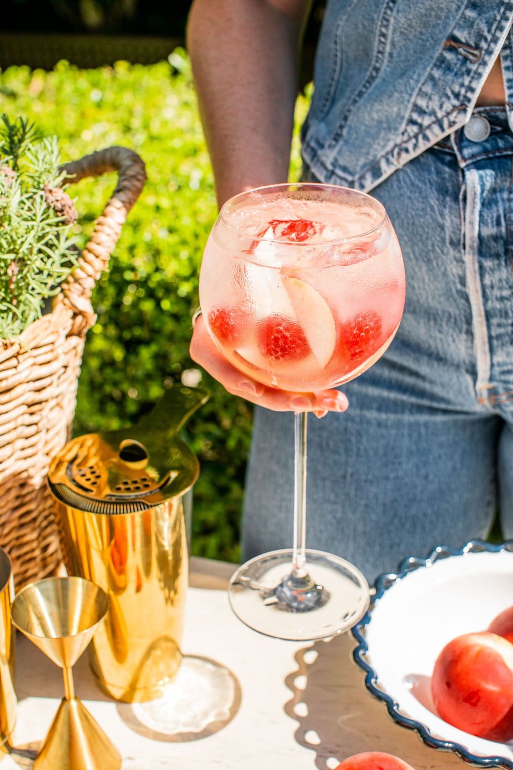 elizabeth dressed in denim holding a wine glass full of peach spritz garnished with raspberries ands lices of peaches