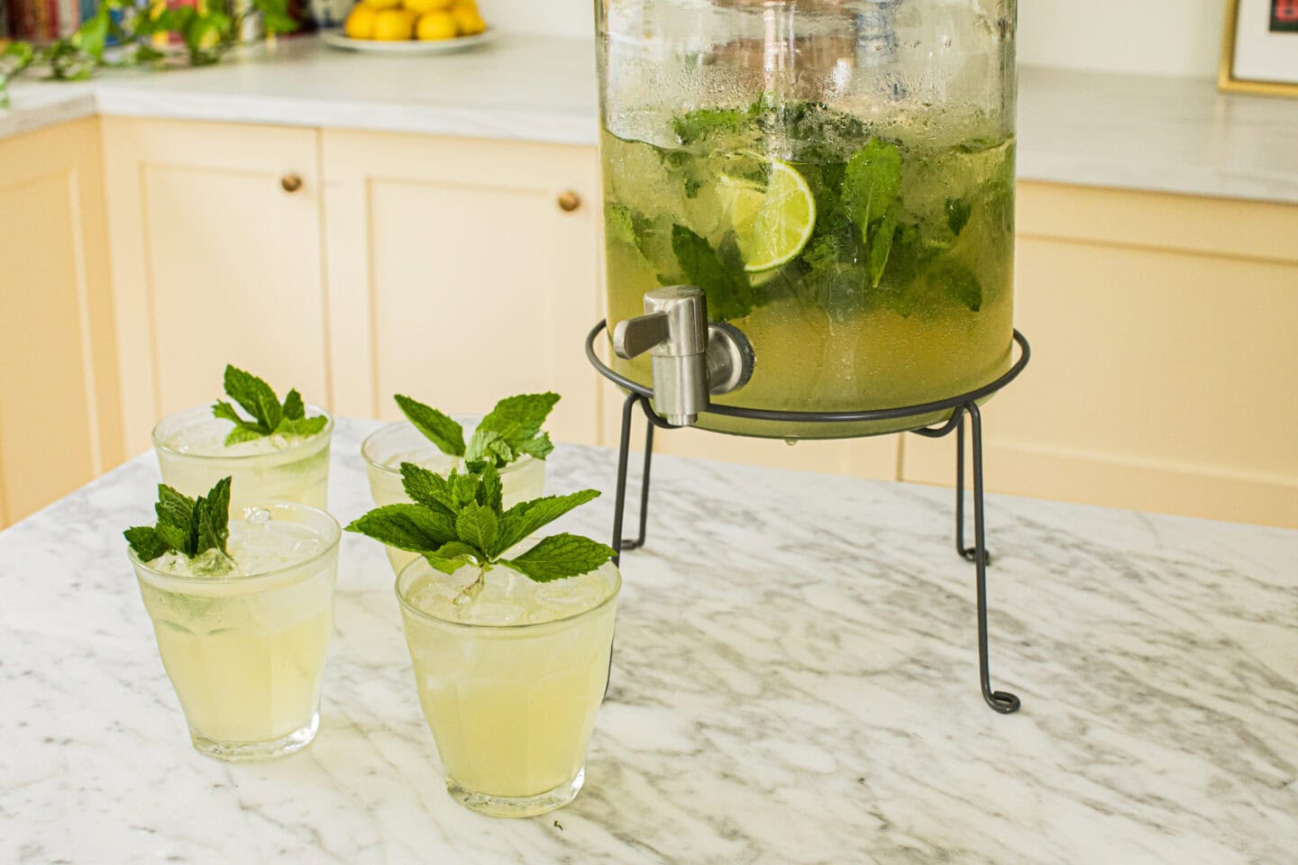 four low glasses filled with hugo spritz and garnished with fresh mint sprits, a drink dispenser half full with hugo spritz