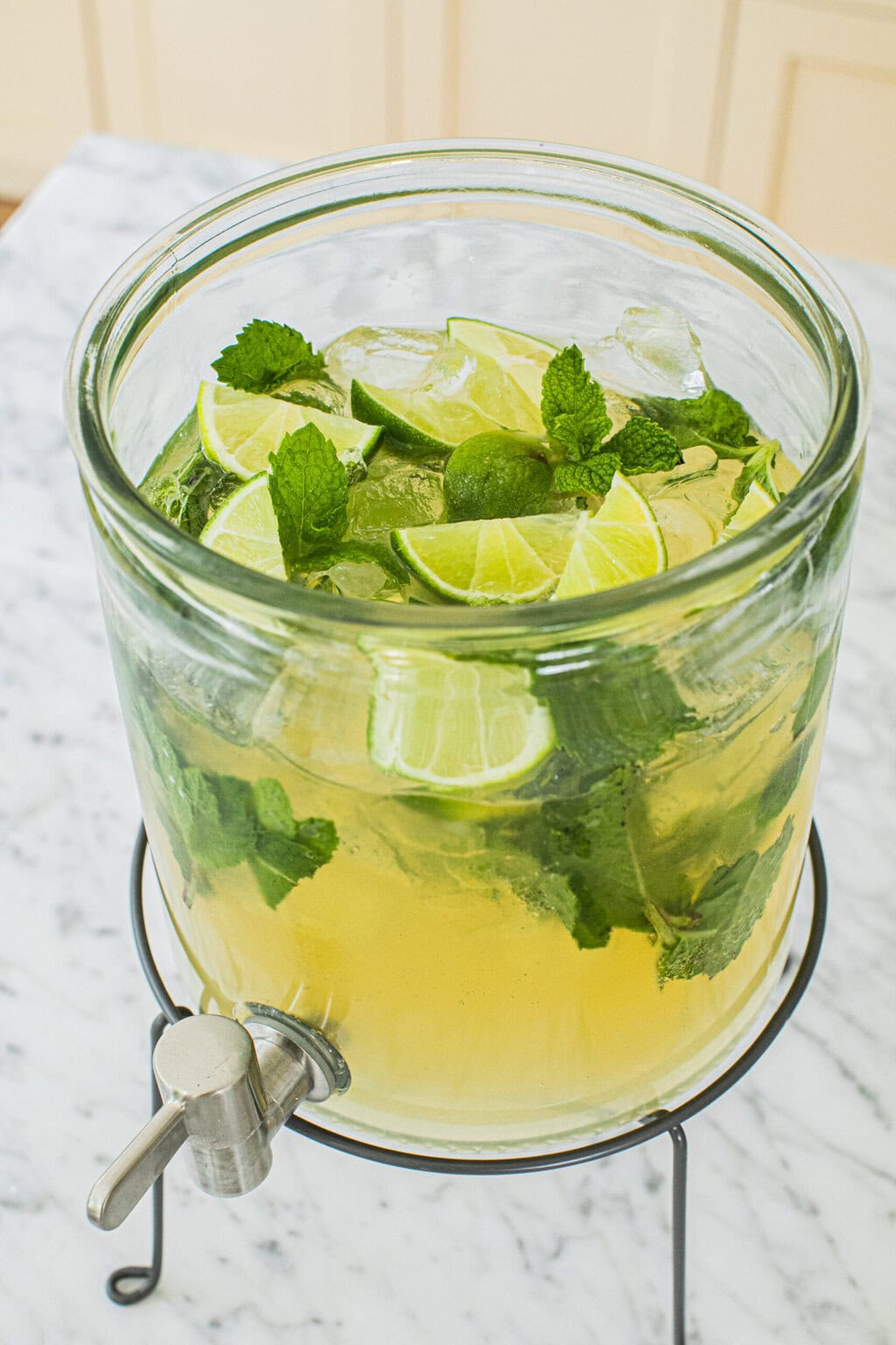 a clear drink dispenser filled with hugo spritz, limes, mint, and ice