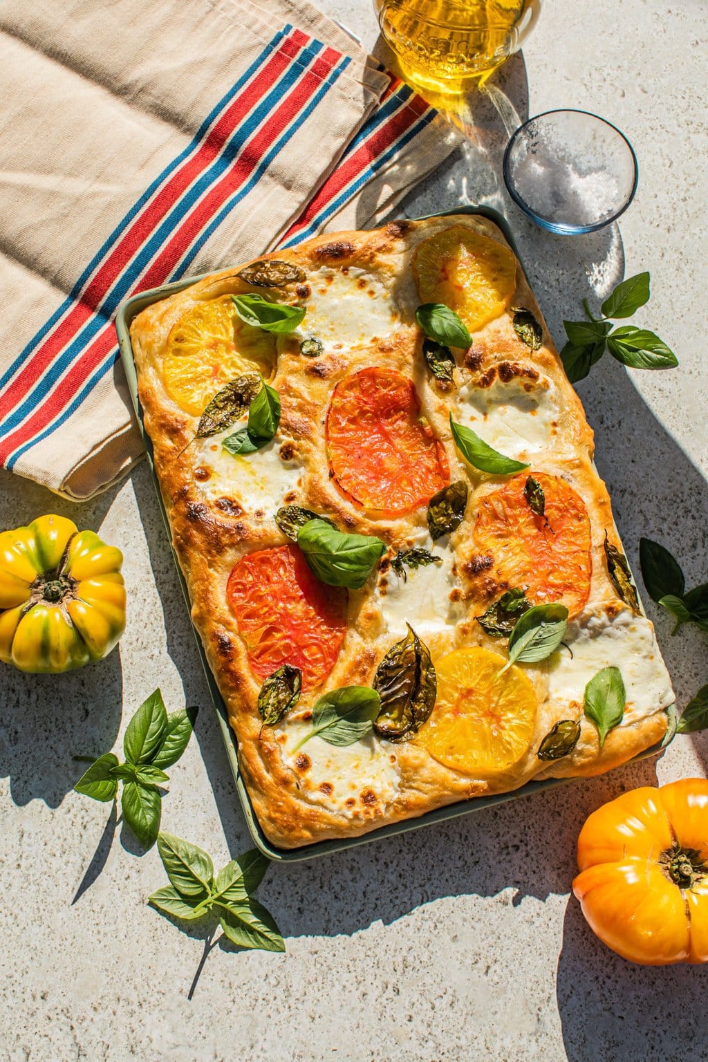 a dish full of baked focaccia topped with caprese ingredients beside excess basil leaves, olives, and a tea towel