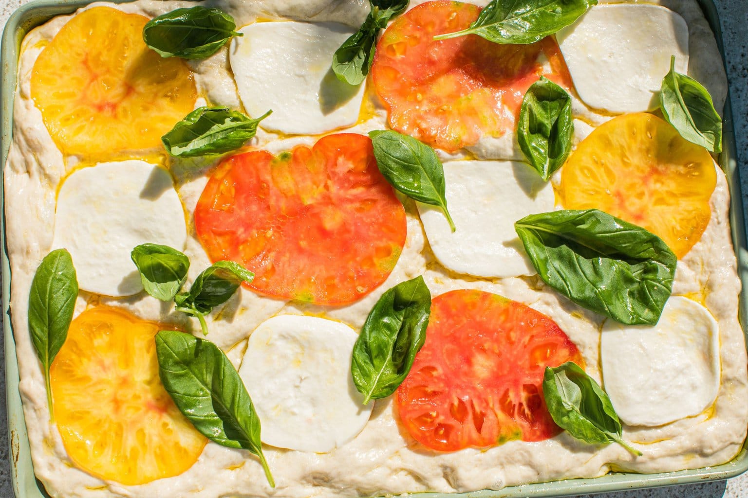 uncooked bread dough topped with thinly sliced tomatoes, mozarella, and basil leaves