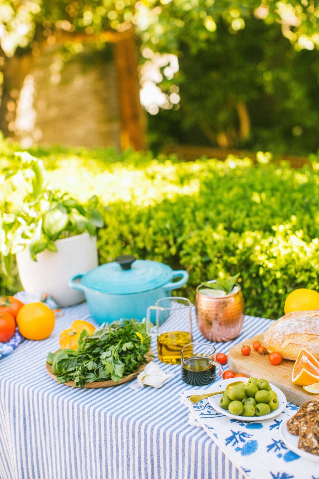 a table with a blue and white striped table cloth topped with fresh herbs, olive oil, balsamic vinegar, olives, tomatoes, drinks, and bread