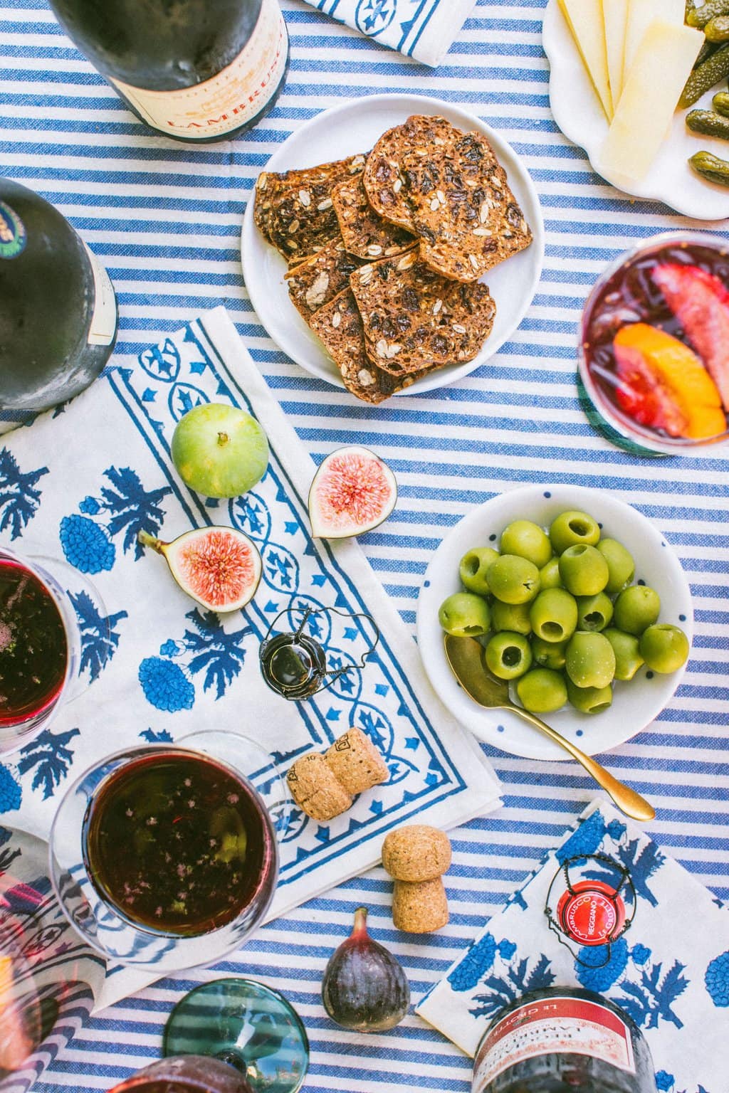 an overhead shot of olives, figs, crackers, and glasses of lambrusco on a white and blue striped table cloth