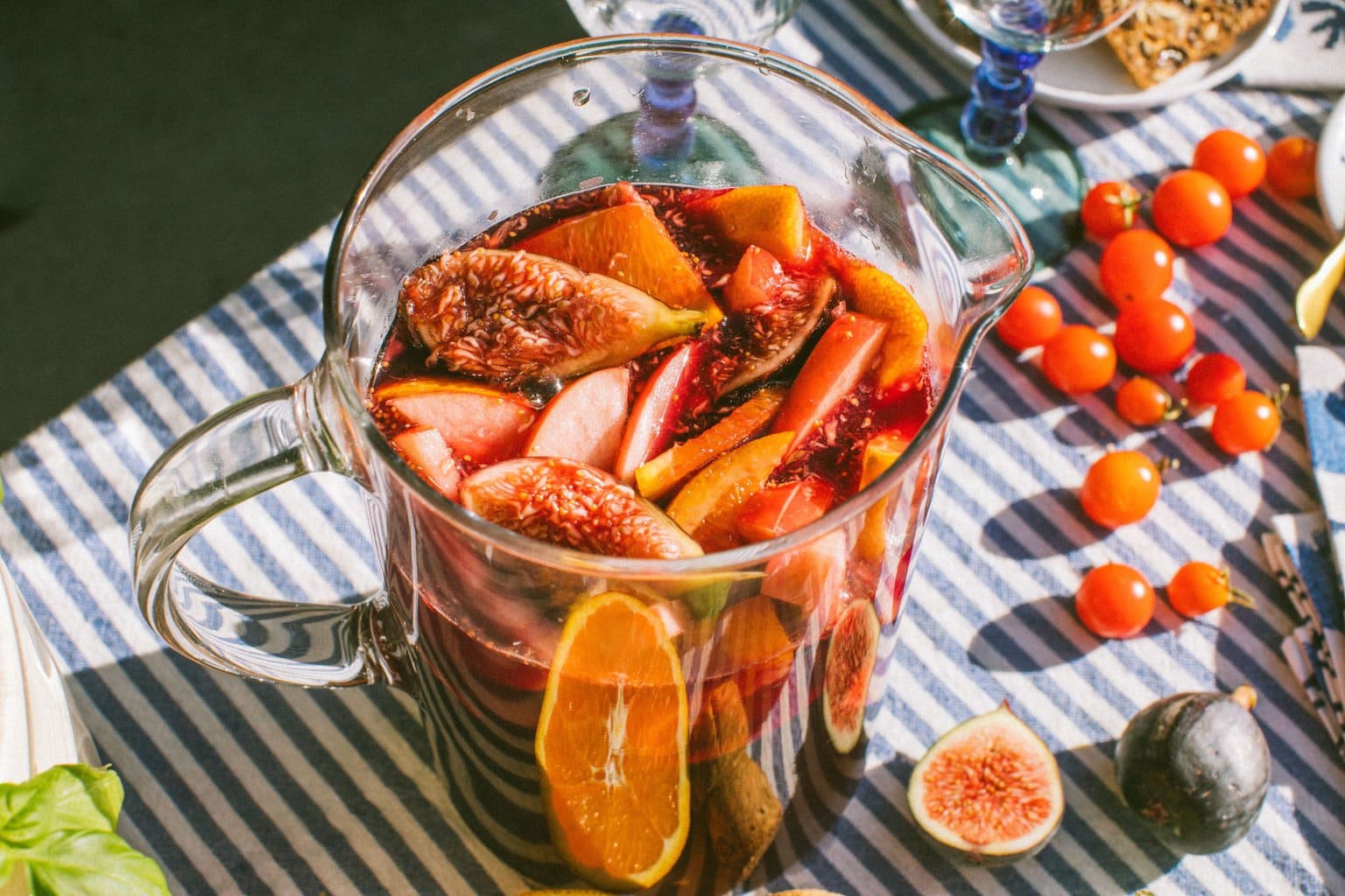 a pitcher of Lambrusco sangria with apple slices, orange slices and fig quarters on a table with a blue stripe table cloth