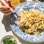 lemon pasta in a blue and white bowl with smaller bowls filled with garnish around it, as well as lemons