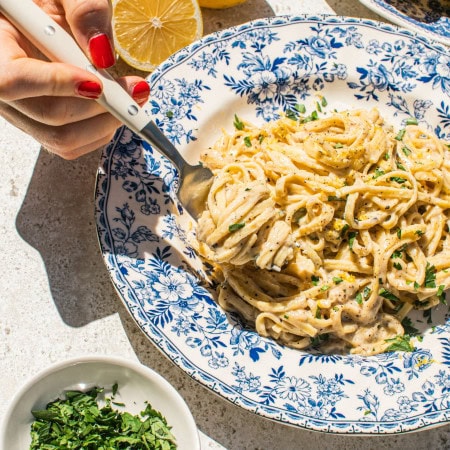 lemon pasta in a blue and white bowl with smaller bowls filled with garnish around it, as well as lemons