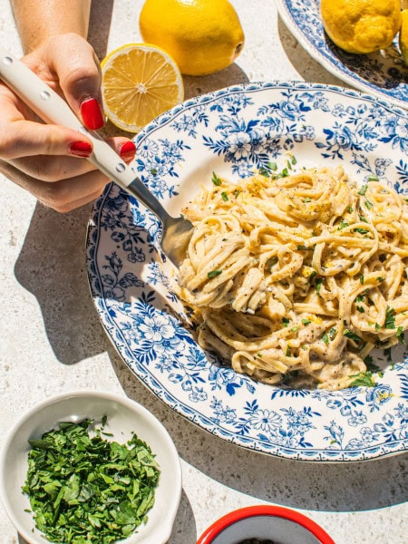 lemon pasta in a blue and white bowl with smaller bowls filled with garnish around it, as well as lemons