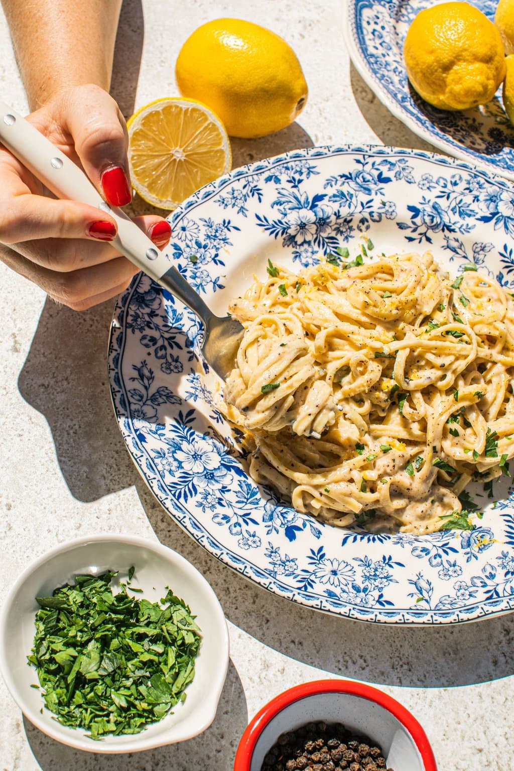 lemon pasta in a blue and white bowl with smaller bowls filled with garnish around it, as well as lemons