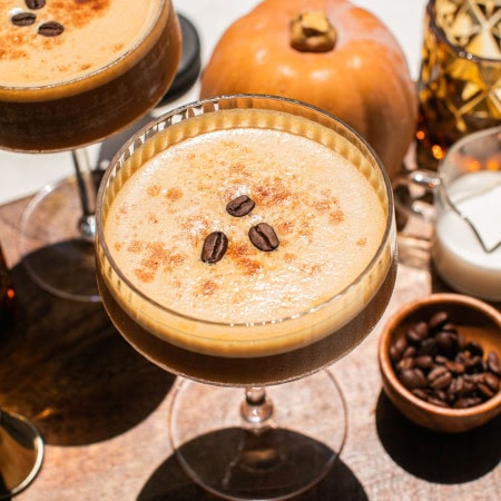 pumpkin spice espresso martini topped with three coffee beans and mini pumpkin decorations seen in the background