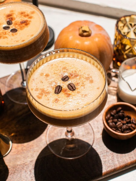 pumpkin spice espresso martini topped with three coffee beans and mini pumpkin decorations seen in the background