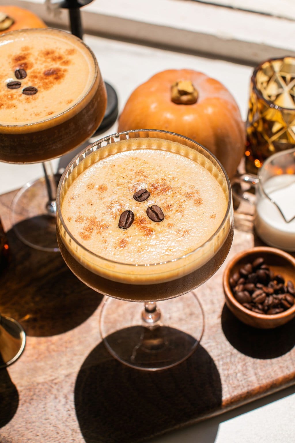 pumpkin spice espresso martini topped with three coffee beans and mini pumpkin decorations seen in the background