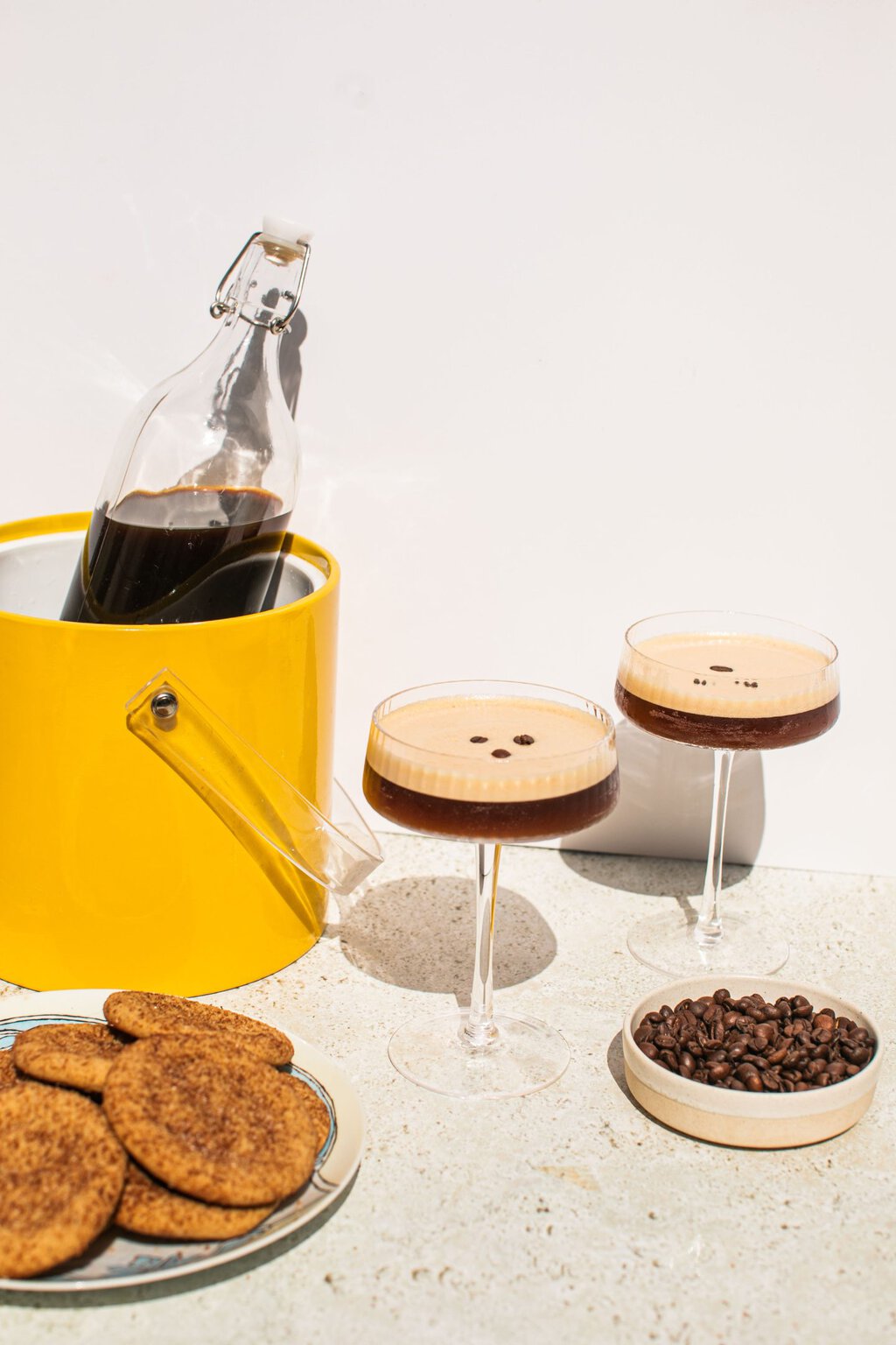a bottle full of espresso martinis with two individual glasses to the side, alongside cookies and espresso beans