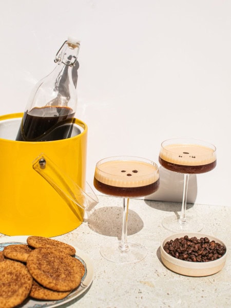 a bottle full of espresso martinis with two individual glasses to the side, alongside cookies and espresso beans