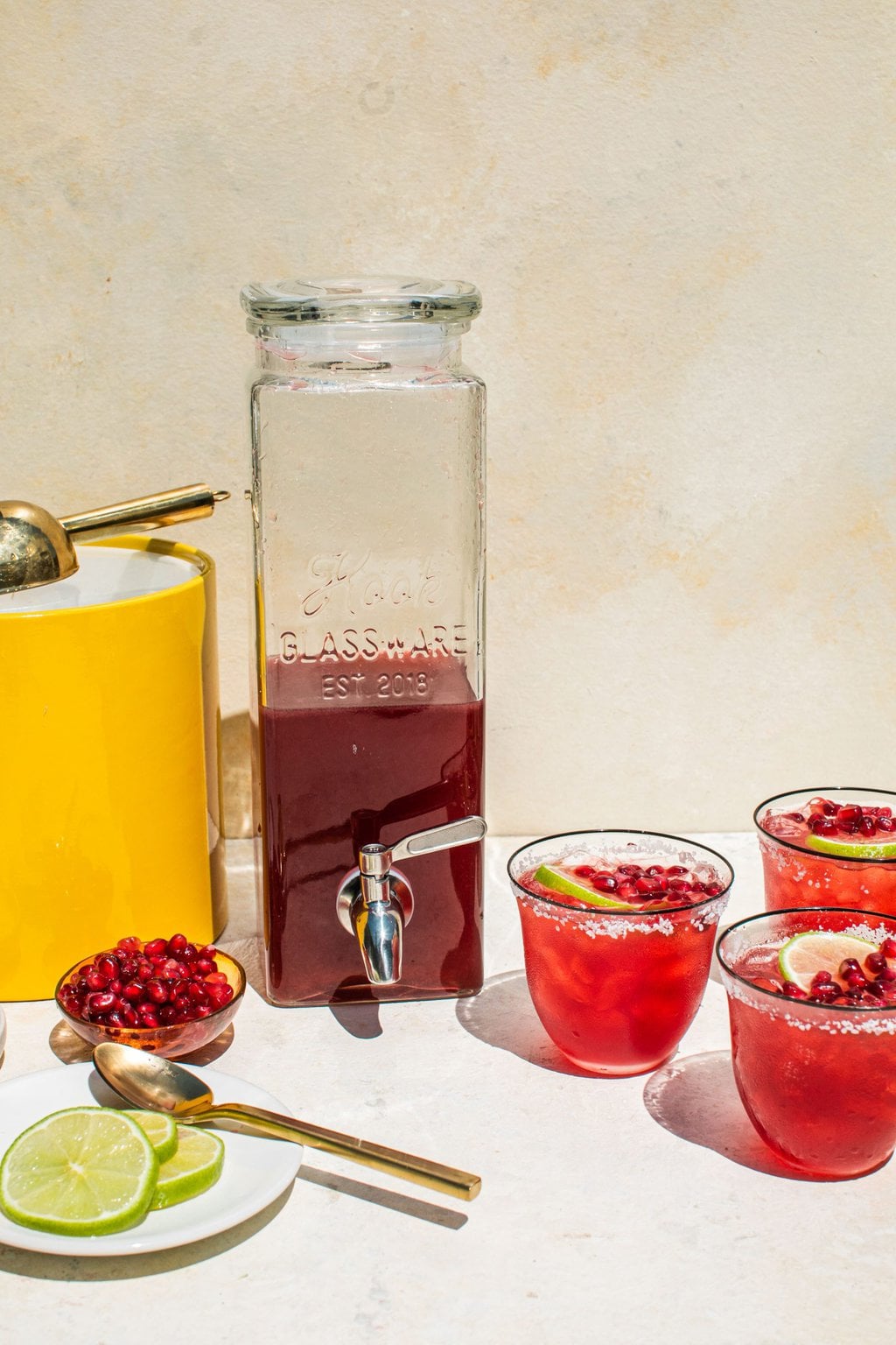 a drink dispenser full of pomegranate margarita liquid and three glasses of pomegranate margaritas with salted rims