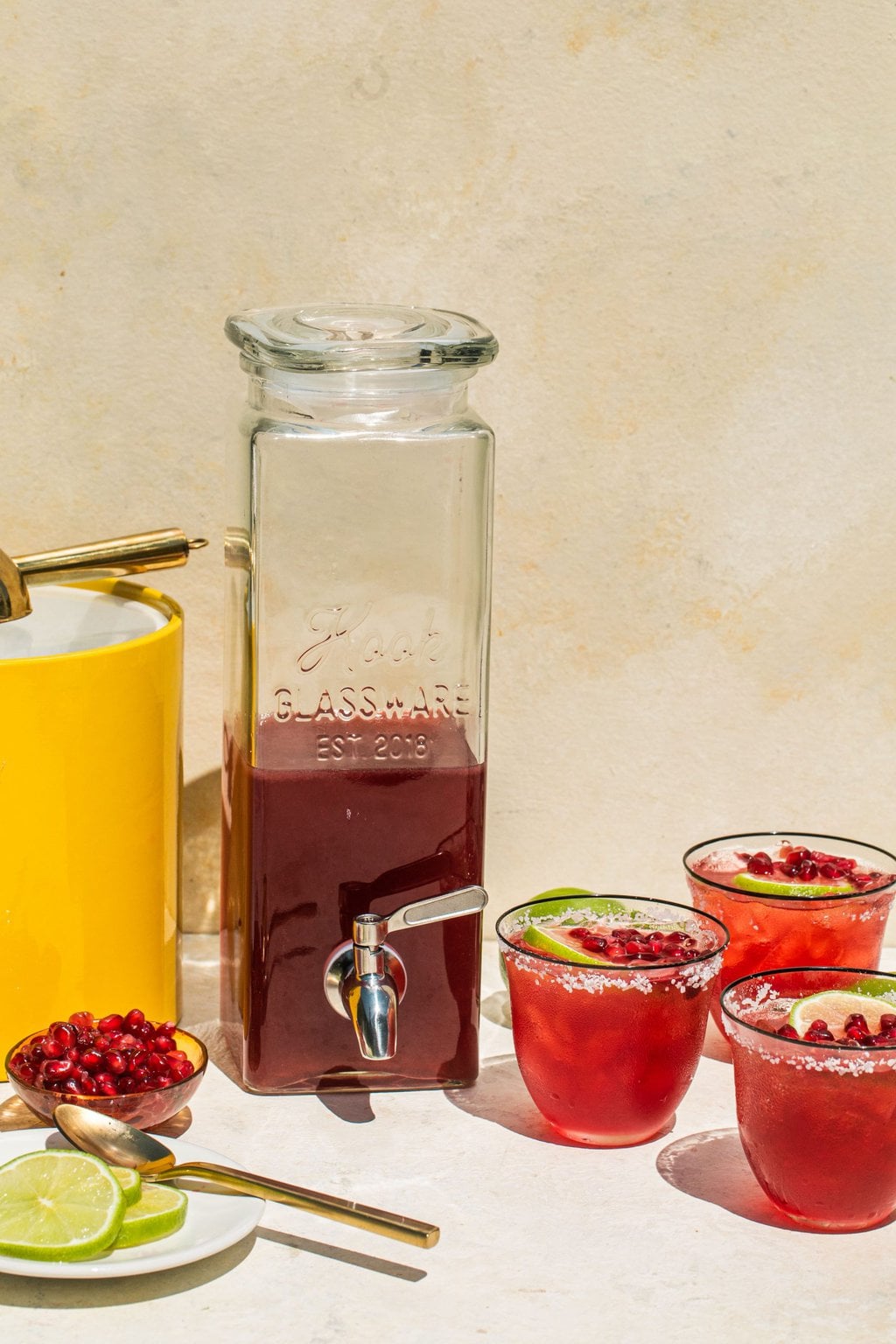 a large dispenser full of pomegranate margaritas beside an ice bucked, pomegranate seeds, and lime slices on the left, with three full glasses on the right