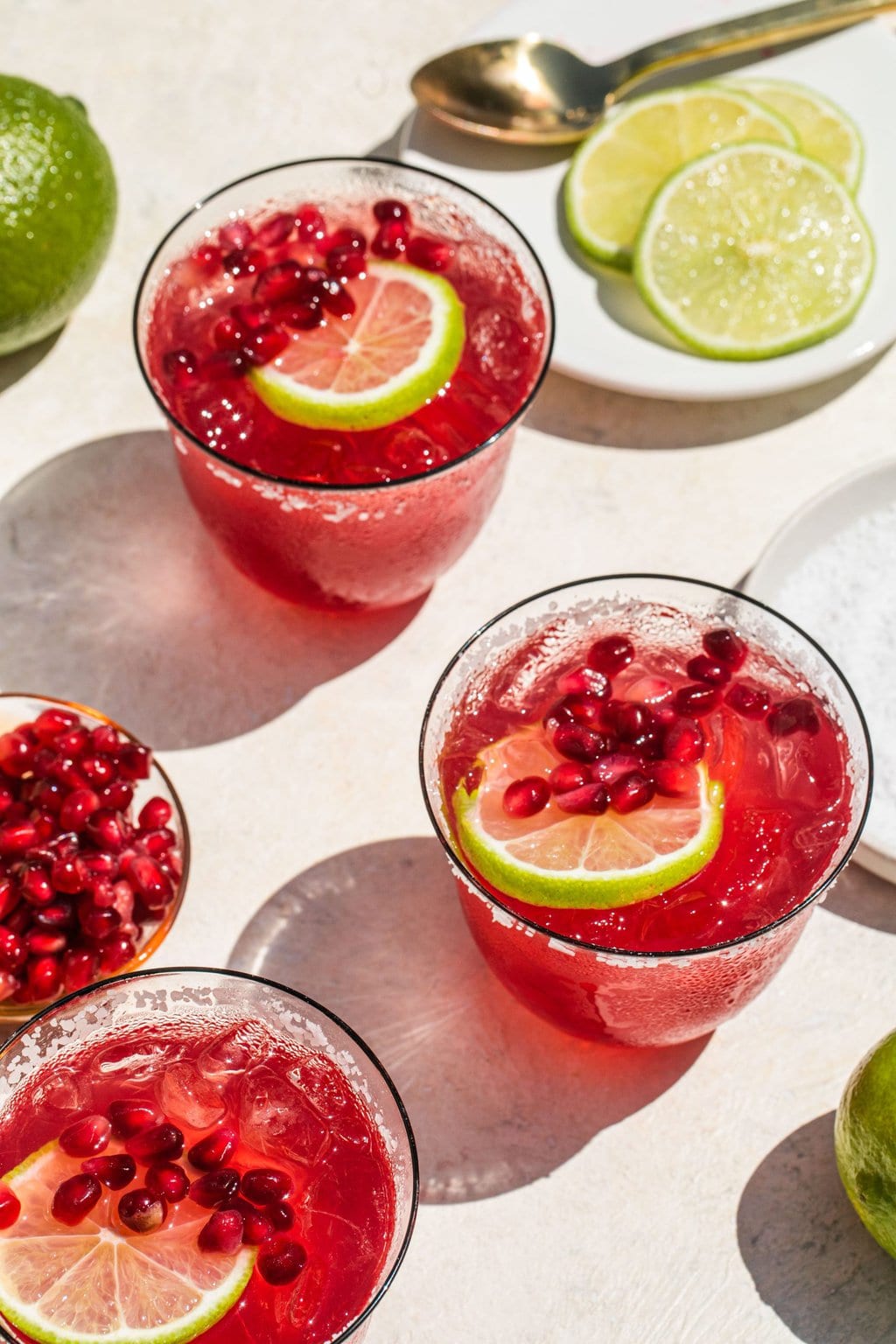 three glasses of pomegranate margaritas garnished with seeds and lime slices besides dishes full of extra garnish