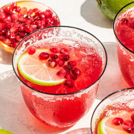 a glass of pomegranate margarita garnished with a lemon slice and pomegranate seeds, along with seeds and a whole lime pictured in the background