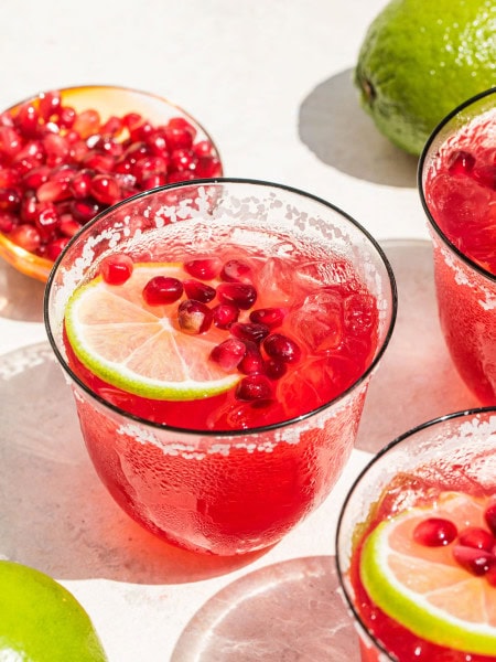 a glass of pomegranate margarita garnished with a lemon slice and pomegranate seeds, along with seeds and a whole lime pictured in the background