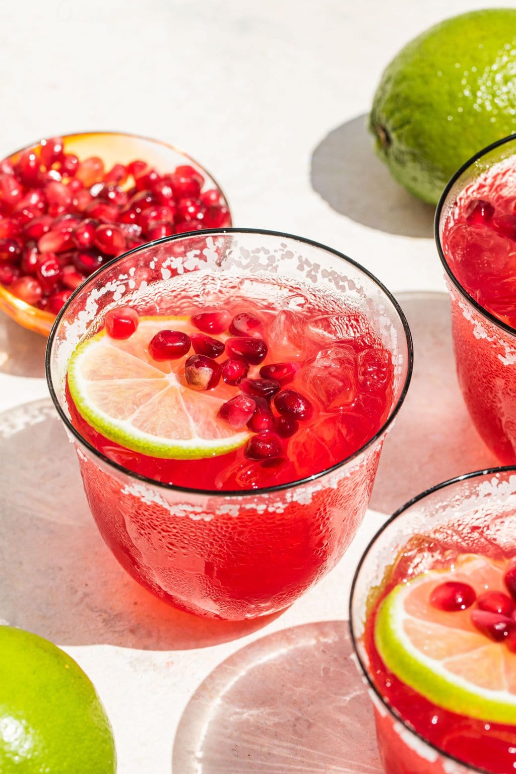 a glass of pomegranate margarita garnished with a lemon slice and pomegranate seeds, along with seeds and a whole lime pictured in the background