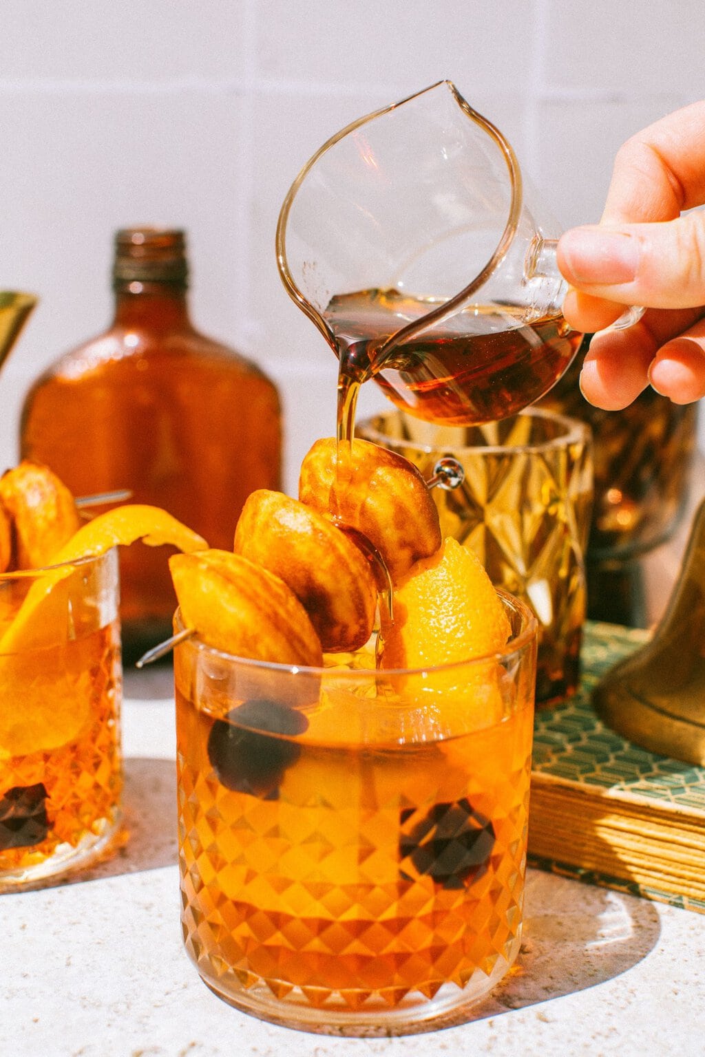 an old fashioned drink with donut hole garnish on top and a hand pouring maple syrup over them