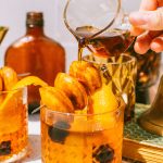 an old fashioned drink with donut hole garnish on top and a hand pouring maple syrup over them