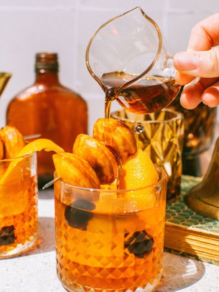 an old fashioned drink with donut hole garnish on top and a hand pouring maple syrup over them