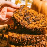 a loaf of pumpkin chocolate chip bread cut into slices and stacked on top of each other, and a hand is lifting the top slice up a bit