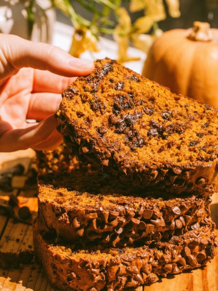 a loaf of pumpkin chocolate chip bread cut into slices and stacked on top of each other, and a hand is lifting the top slice up a bit