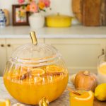 glass pumpkin full of punch with a glass in front of it and mini pumpkins scattered around