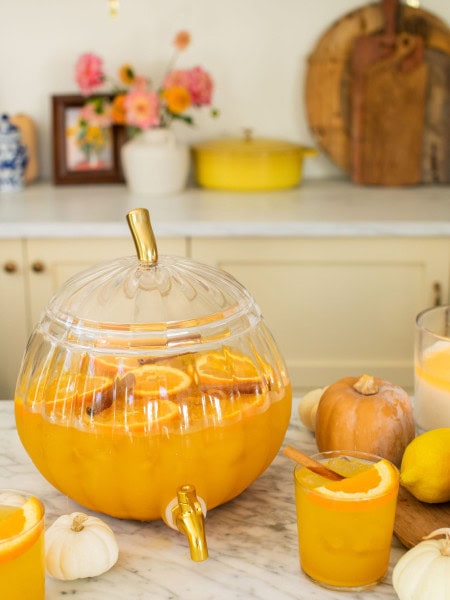 glass pumpkin full of punch with a glass in front of it and mini pumpkins scattered around