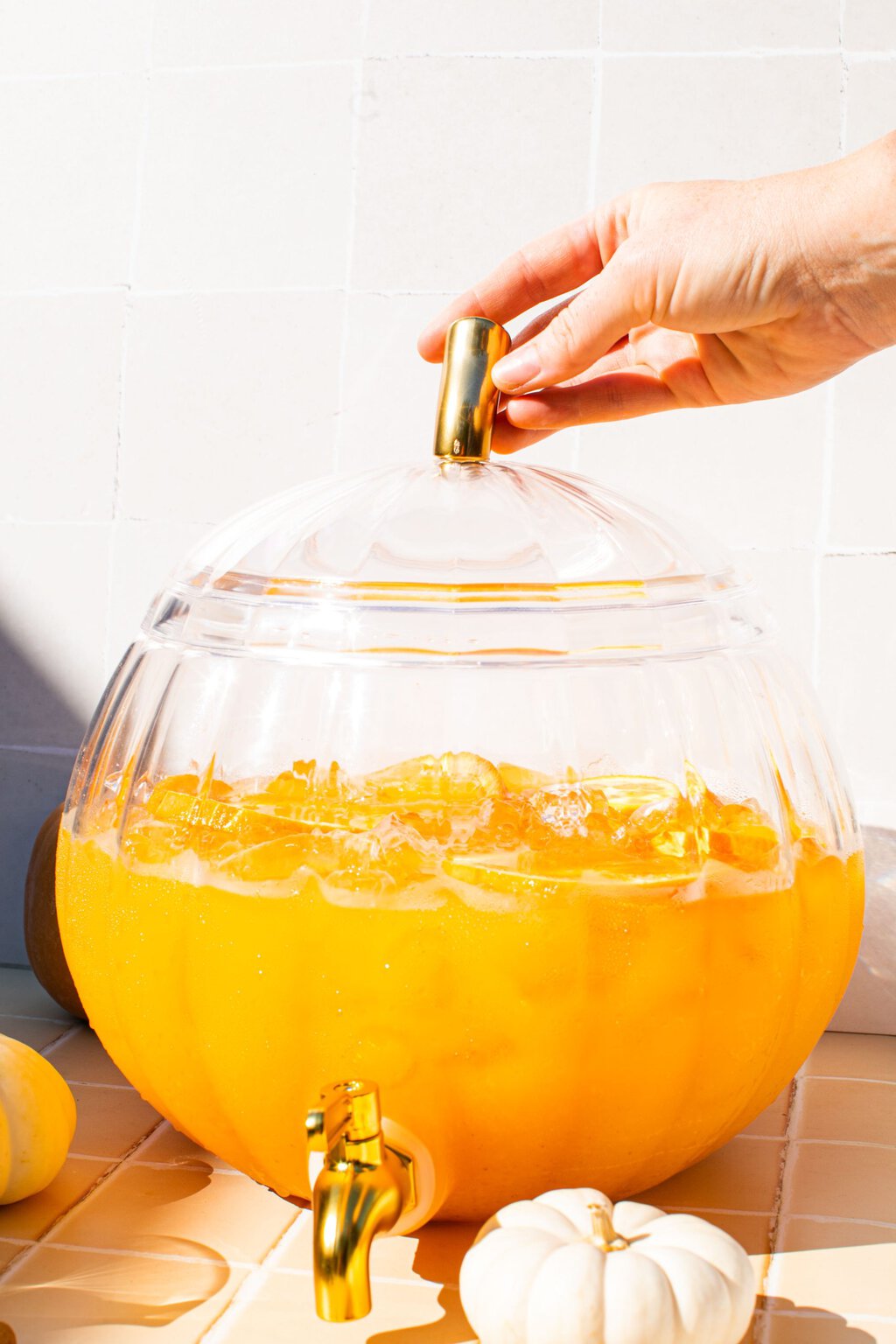 a hand on top of the glass pumpkin which is holding spiked pumpkin punch and has mini pumpkins around it