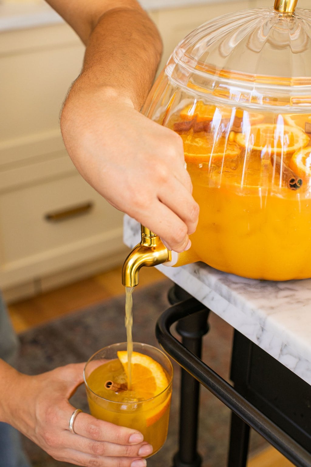 a hand dispensing pumpkin punch from the large glass pitcher into a cocktail glass garnished with orange and cinnamon sticks