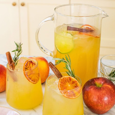 a large pitcher of apple cider margaritas in the background with two garnished glasses in the front alongside whole apples