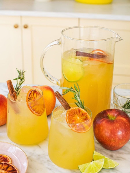 a large pitcher of apple cider margaritas in the background with two garnished glasses in the front alongside whole apples