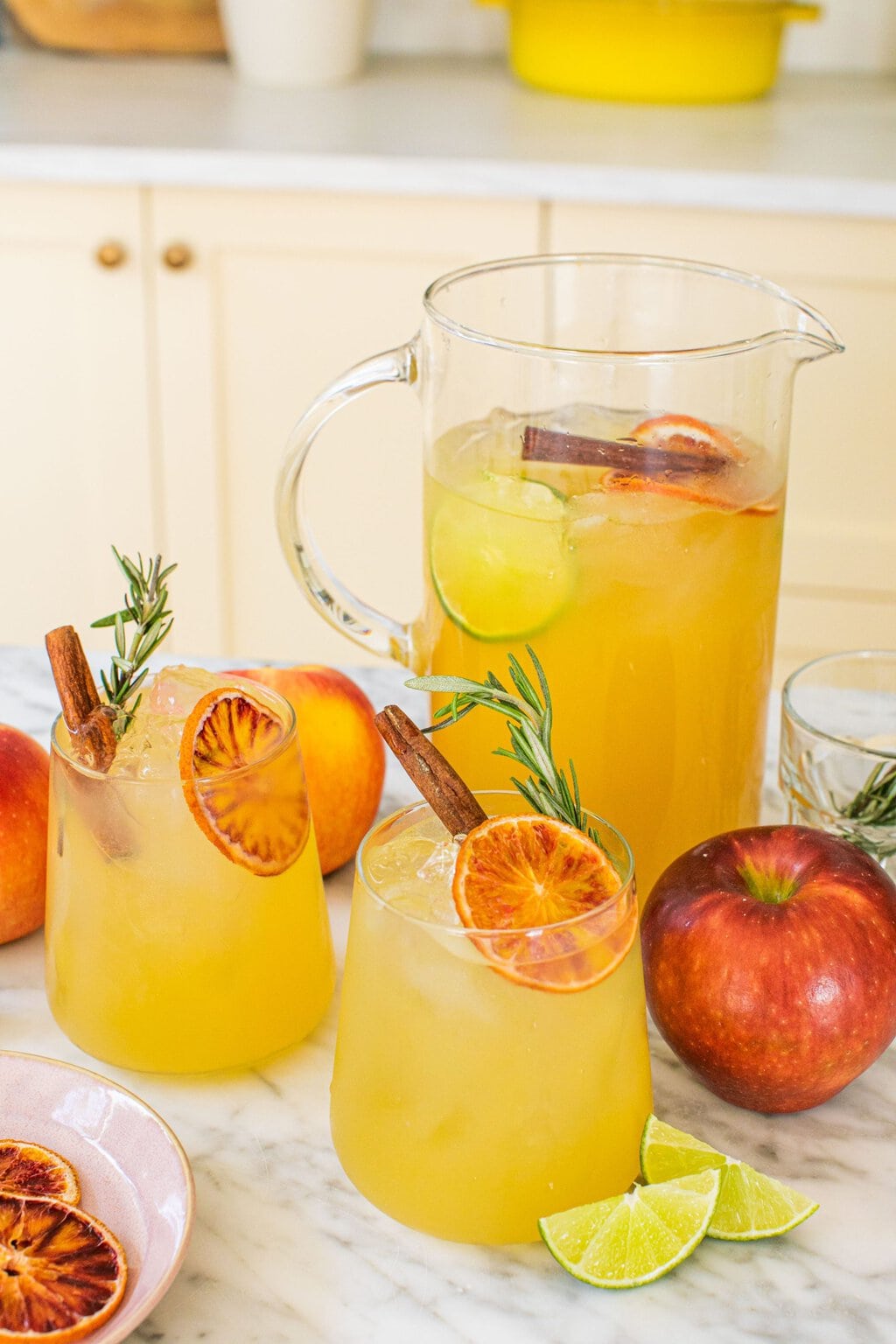 a large pitcher of apple cider margaritas in the background with two garnished glasses in the front alongside whole apples