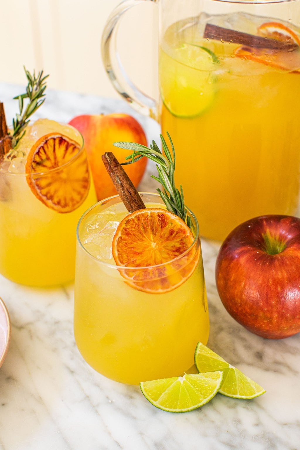 a large pitcher of an apple cider margarita in the background with two garnished glasses in front beside fresh apples