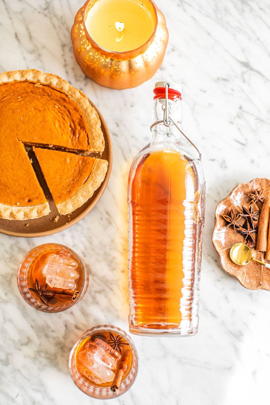 an overhead shot of two glasses of old fashioneds, a large bottle of the cocktail, a pumpkin pie, and garnish