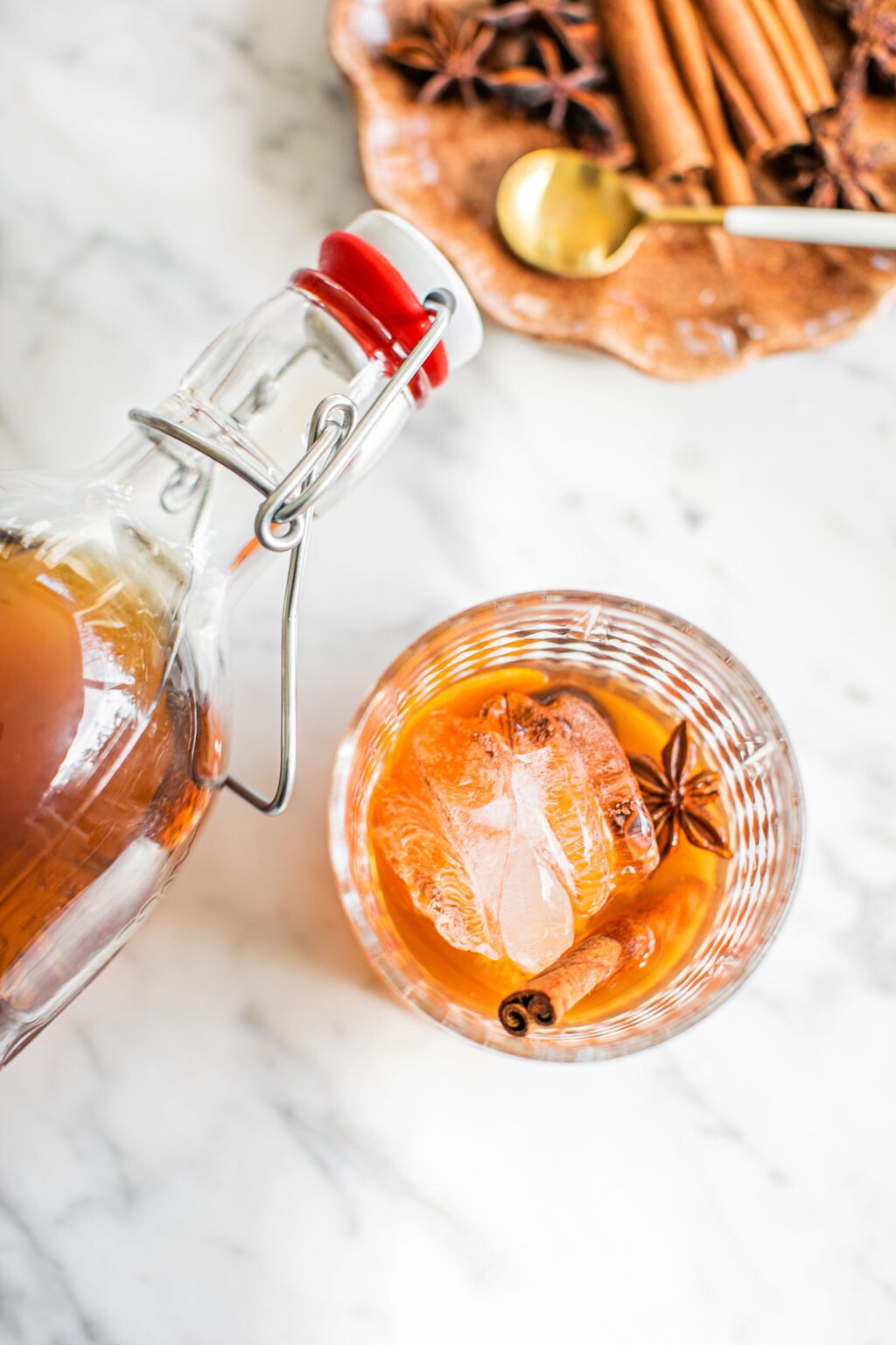 an overhead photo of an old fashioned in a lowball glass with star anise and a cinnamon stick as garnish, and a swing top bottle over it