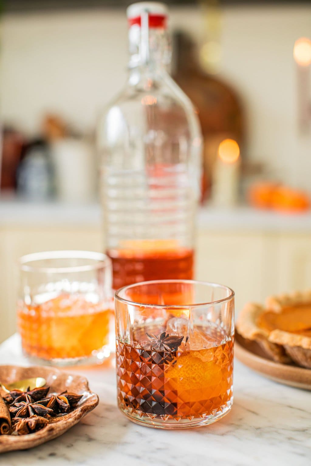 two glasses of old fashioneds beside a plate with garnish on it and a large bottle full of the cocktail and a pumpkin pie out of focus in the background